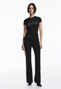Crna uska majica s natpisom 'Party Call' u srebrnom rukopisu, spojena s crnim hlačama širokih nogavica s detaljem pertle i okruglim metalnim ukrasom.