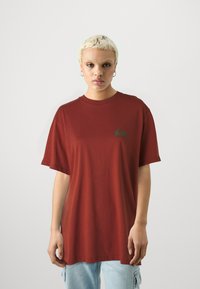 Preveľký marhuľový tričko vyrobené z mäkkého bavlneného materiálu. Má malé zelené logo na ľavej strane hrude a krátke rukávy. Klasický design s okrúhlym výstrihom.