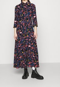 Lang, marineblauwe jurk met een multicolore abstracte luipaardprint, knopen aan de voorkant en driekwart mouwen. Gecombineerd met zwarte enkelboots.
