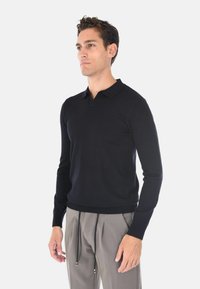Polo shirt nera a maniche lunghe in maglia con orlo e colletto a costine, abbinata a pantaloni plissettati di colore chiaro. Dettaglio della vestibilità rilassata visibile.