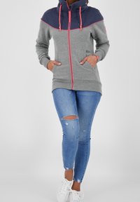 Felpa grigia e blu navy con cerniera e accenti rosa, dotata di un alto colletto e tasche laterali, abbinata a jeans blu scoloriti e sneakers bianche.