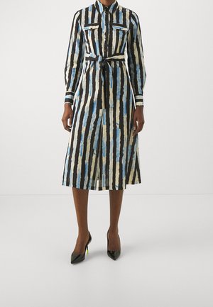 Femme portant une robe chemise à col avec manches longues, rayures verticales noires, bleues et beiges, une ceinture à la taille et des talons hauts noirs.