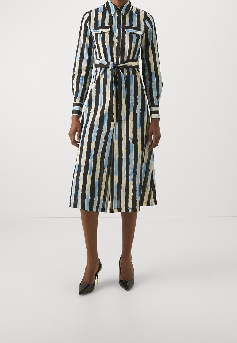 Femme portant une robe chemise à col avec manches longues, rayures verticales noires, bleues et beiges, une ceinture à la taille et des talons hauts noirs.