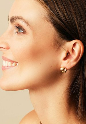 Lächelnde Frau mit braunen Haaren präsentiert goldene, knotenförmige Ohrringe, im Close-up-Profil vor einem neutralen beigen Hintergrund.