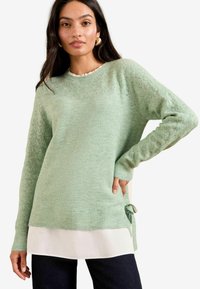 Hellgrüner Strickpullover mit Lochmuster, gerippten Bündchen und Saum sowie einer seitlichen Krawatte. Darüber trägt er ein weißes, geschichtetes Hemd.