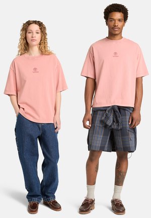 Twee modellen dragen bijpassende roze Timberland T-shirts; de ene combineert het met losse jeans, de ander met geruite shorts en een shirt om de taille geknoopt.