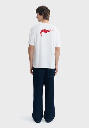 Muškarac stoji okrenut leđima, nosi bijelu majicu s crvenim motivom škampi na leđima i tamne široke hlače.