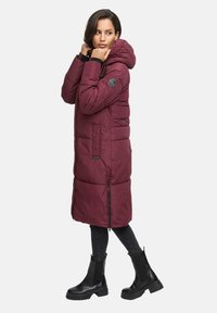 Woman adjusting hood on long burgundy quilted coat, wearing black leggings and black chunky boots, standing against white background.