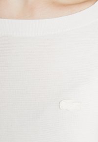 Vit stickad tröja med rund halsringning och ett subtilt strukturerat mönster. Framsidan har en liten, vit broderad logotyp på vänster bröst.