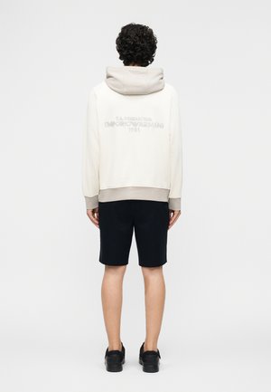 Άτομο με σγουρά μαλλιά που φοράει λευκή φούτερ με μπεζ κουκούλα και μανσέτες, μαύρο σορτς και μαύρα παπούτσια, όρθιο με την πλάτη προς τα εμπρός.