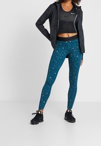 Leggings verde azulado con estampado estrellado, combinados con una camiseta de tirantes negra y un cortavientos. Zapatos deportivos negros complementan el conjunto.