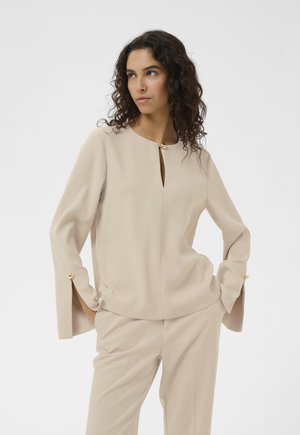 Femme aux cheveux bouclés portant un chemisier beige à manches longues avec des boutons dorés sur les poignets et le col, ainsi qu'un pantalon beige assorti.