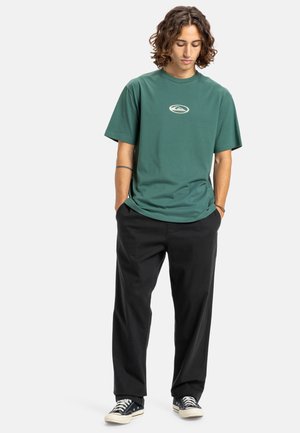 Green cotton t-shirt with a round neckline and short sleeves, featuring a small logo in the center; paired with black trousers and sneakers.