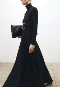 Schwarzes langärmliges Kleid mit hohem Kragen, das einen figurbetonten Oberteil und einen fließenden Rock hat. Kombiniert mit einer strukturierten Clutch und kreisförmigem Akzent.