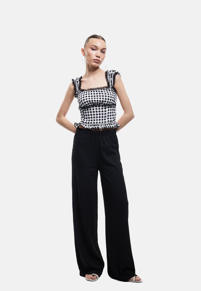 Geruite zwart-witte top met ruches aan de mouwen en zwarte accenten, gecombineerd met hoge taille zwarte wijde broek en lichte sandalen.