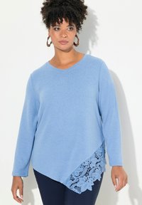 Maglietta blu a maniche lunghe con scollo a V, caratterizzata da un accento in pizzo testurizzato lungo l'orlo asimmetrico, abbinata a pantaloni scuri.