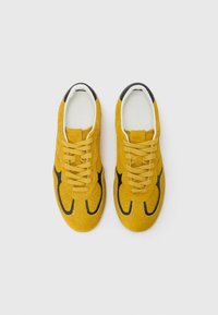 Gelbveloursleder-Schnürsneaker mit schwarzen Akzenten, die über durchbrochene Elemente und ein weißes Innenfutter verfügen und ein abgerundetes Fußdesign zeigen.