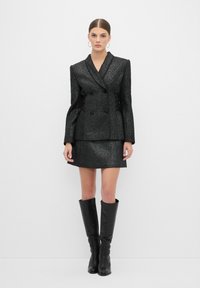 Blazer nero brillante doppiopetto con spalle larghe, abbinato a una mini gonna coordinata e stivali neri sopra il ginocchio.
