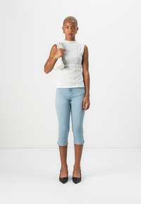 Hvit ermeløs topp med strukturert bølge mønster, lyseblå avkuttede jeans, og svarte spisse hæler. Holder en liten hvit håndveske.