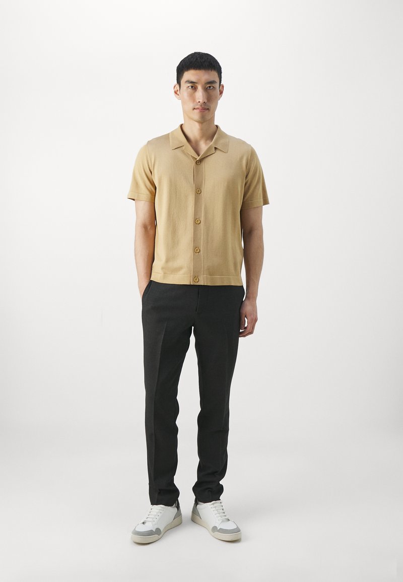 Beige Kurzarm-Hemd mit Knöpfen und Kragen, kombiniert mit schwarzen Hosen und weißen Sneakers. Glatte Textur, einfaches Design, neutrale Farben.