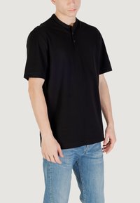 Camisa polo negra de manga corta con cuello abotonado, confeccionada en tela suave, exhibida en un modelo que lleva jeans de color azul claro.