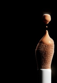 Pennello per trucco con setole morbide di un caldo marrone chiaro, con una goccia di fondotinta liquido in una tonalità nude calda, su uno sfondo nero.