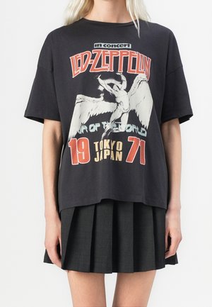 Personne portant un t-shirt de concert Led Zeppelin noir avec une image d'une figure ailée, le texte "19 Tokyo Japan 71" et une jupe plissée sombre.