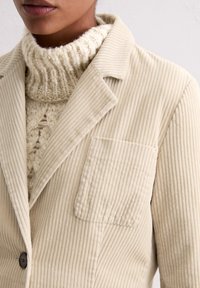 Beige corduroy blazer met een omgeslagen kraag, een enkele knoopsluiting en een borstzak. De textuur is geribbeld en zacht. Gehaakt crème-coloured coltrui eronder.