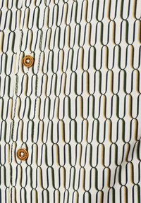 Korte mouwen shirt met een witte basis, met een geometrisch patroon in groen, geel en zwart. Inclusief een bruine knoop en zichtbare stiksels.