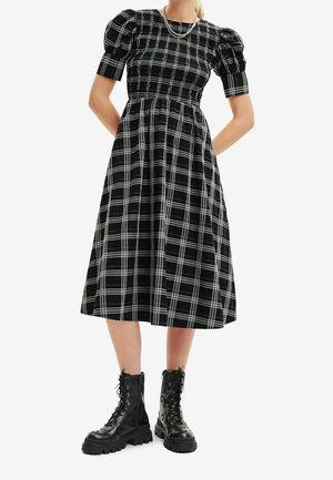 Woman wearing a black and white checkered midi dress with puff sleeves and black chunky lace-up boots, standing with hands behind back.