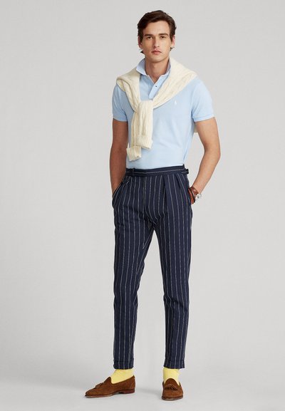 Mand iført lyseblå polo, cremefarvet sweater draperet over skuldrene, marineblå bukser med nålestriber, gule sokker og brune loafers med kvaster, stående med hænderne i lommerne.