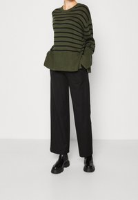 Khaki gestreifter Pullover mit geripptem Saum, kombiniert mit schwarzer weit geschnittener Hose und klobigen schwarzen Knöchelschuhen. Weiche Stricktextur.