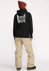 Haut à capuche noir avec un motif zèbre au dos, associé à un pantalon cargo beige et un bonnet en tricot à motifs. Les bottes sont dotées d'une semelle en caoutchouc.