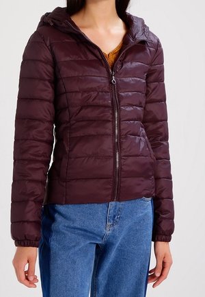 Femme portant une doudoune sombre violette à fermeture éclair avec capuche et un jean bleu, debout devant un fond clair uni.