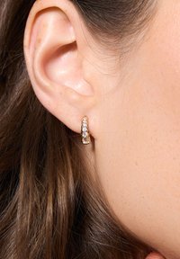 Złota kolczyk w kształcie pierścienia z pięcioma przezroczystymi kamieniami, o gładkiej teksturze i wypolerowanej powierzchni, noszony przez modelkę z długimi, brązowymi włosami.