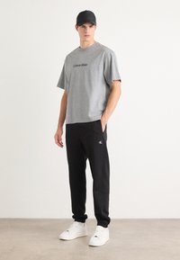 Grå bomull T-skjorte med "Calvin Klein"-logo, kombinert med svarte joggebukser med hvit logo. Modell har på seg hvite sneakers og en svart caps.