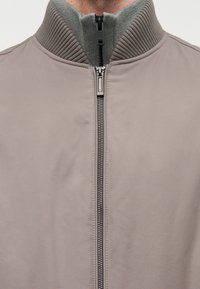 Ανοιχτό γκρι bomber μπουφάν με υψηλό ριμπ γιακά. Διαθέτει μαύρο φερμουάρ κλεισίματος και λεία υφή υφάσματος, κατάλληλο για casual ντύσιμο.