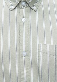 Πράσινο ριγέ πουκάμισο με στενές λευκές και σκούρες ρίγες, γιακά με κουμπιά, εμπρόσθια πατιλέτα με ανοιχτόχρωμα κουμπιά και τσέπη στο στήθος.