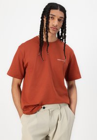 Tricou scurt de culoare ruginită din bumbac, cu un detaliu al logo-ului alb pe piept, asortat cu pantaloni bej deschis.