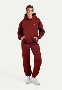 Conjunto de sudadera con capucha burdeos y pantalones deportivos a juego, confeccionado con tela suave. Cuenta con un bolsillo delantero y puños elásticos. Estilizado con zapatillas blancas.