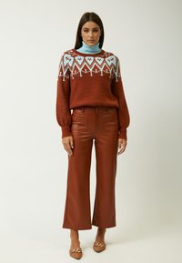 Rostfarbener Strickpullover mit hellblauen geometrischen Mustern, kombiniert mit braunen, weiten Hosen aus Kunstleder und spitz zulaufenden Schuhen.