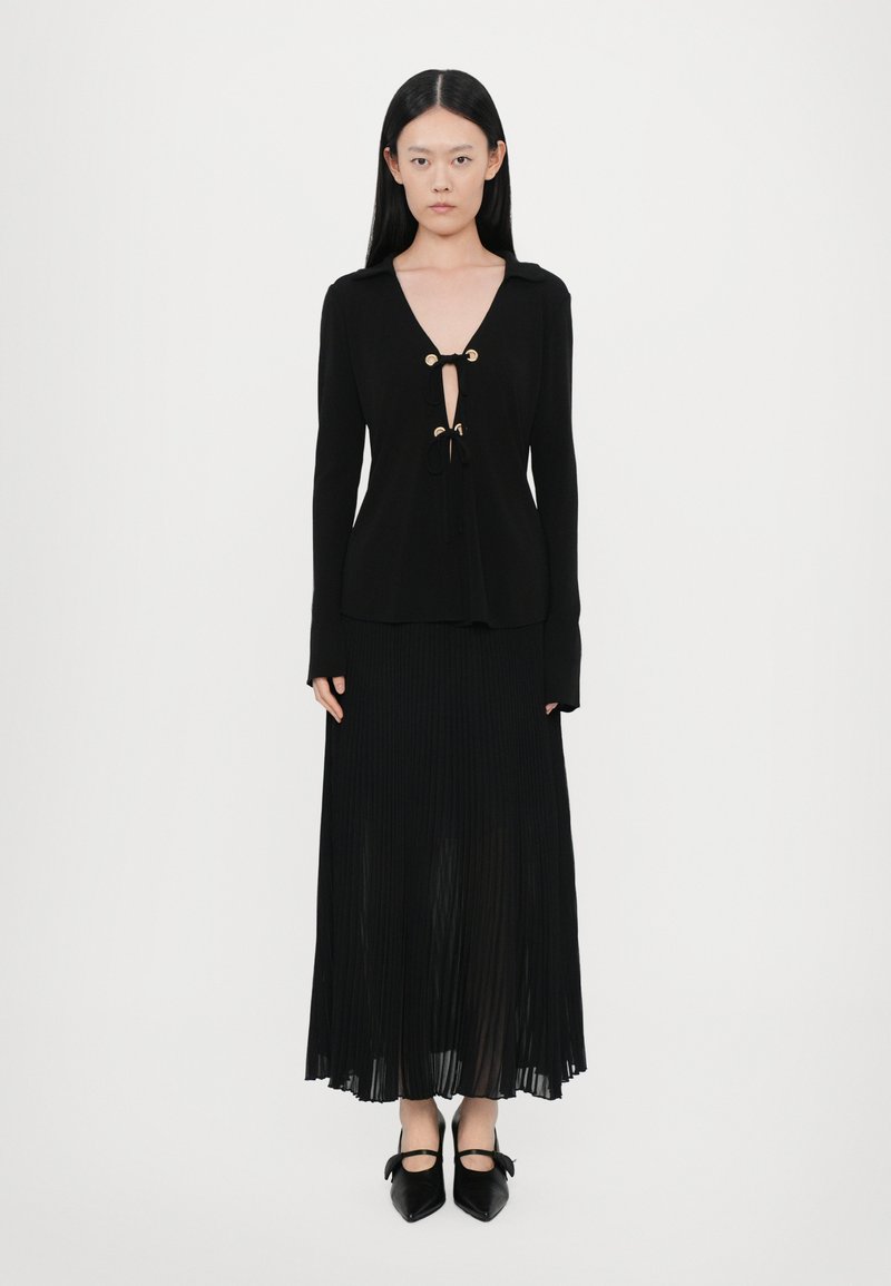Black long-sleeve top with a V-neck and gold toggle accents, paired with a pleated black skirt and pointed black shoes.