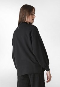 Svart oversized sweatshirt med rundad hals, långa ärmar och en subtil logotyp på baksidan. Mjuk bomullstyg med matt yta.