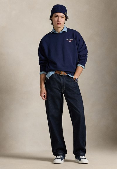 Granatowa bluza z kapturem z logo "POLO SPORT", pod spodem jasnoniebieska koszula jeansowa, ciemne, luźne dżinsy oraz białe trampki. Brązowy pasek i niebieska czapka beanie.