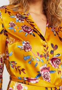 Gul satinblus med blommönster i röda och blå nyanser, en krage och framknappar, accentuerad med knyt i midjan.
