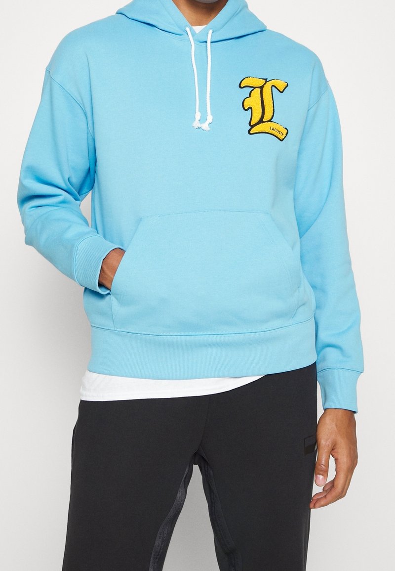 Homme portant un sweat à capuche bleu clair avec un patch jaune "L" sur la poitrine, des cordons blancs, une poche avant et un pantalon noir.