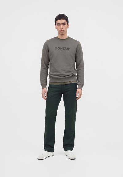 Млад мъж, облечен със зелен суичър DONDUP, тъмнозелени панталони и бели маратонки на светъл едноцветен фон.