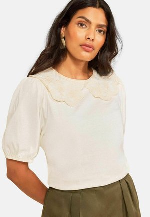 Hosszú, sötét hajú nő krémszínű blúzt visel hímzett hullámos gallérral és puffos rövid ujjakkal, amit olivazöld nadrággal párosít.