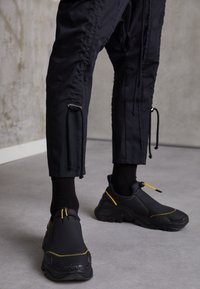 Schwarze Slip-On-Sneaker mit gelben Akzenten, strukturiertem Gummisohle und glattem Obermaterial. Die Hose hat eine lockere Passform und Kordelzugdetails.