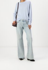 Person wearing light blue sweater, wide-leg faded jeans, black shoes, and carrying a small dark brown shoulder bag against a white background.